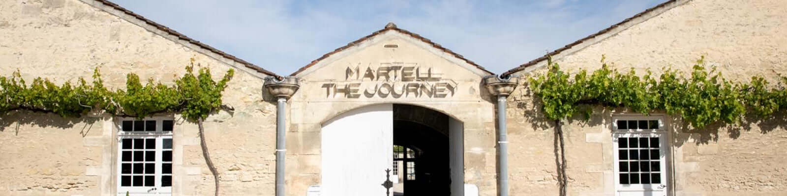 Facade of a historic stone building with green vines and two windows. The entrance features an arched sign that reads Martell The Journey.