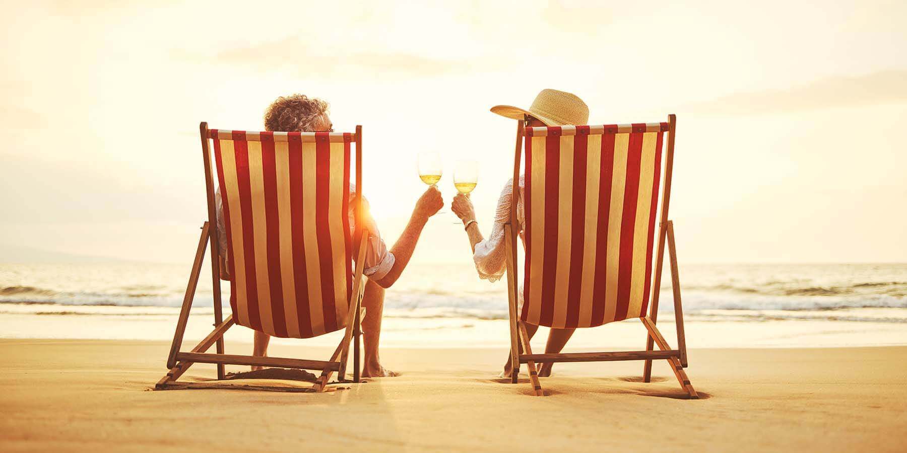 Two people sit in red-and-white striped beach chairs, facing the ocean at sunset. They hold wine glasses and toast, with the sun setting on the horizon creating a warm glow. One person wears a wide-brimmed hat. The scene is peaceful and idyllic.