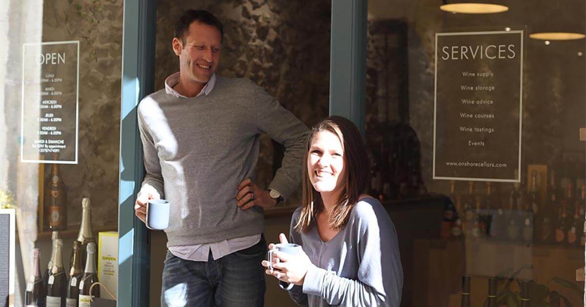 A man and a woman are standing outside a store with blue doors labeled SERVICES. They are both holding mugs and smiling in the sunlight. Behind them, there are wine bottles displayed near the entrance.