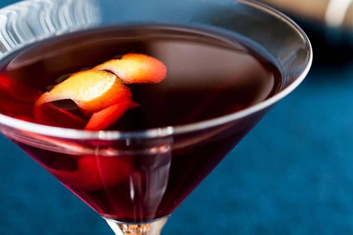 A close-up of a clear martini glass filled with a dark red cocktail, garnished with a twist of orange peel. The background is softly blurred and has a deep blue hue.