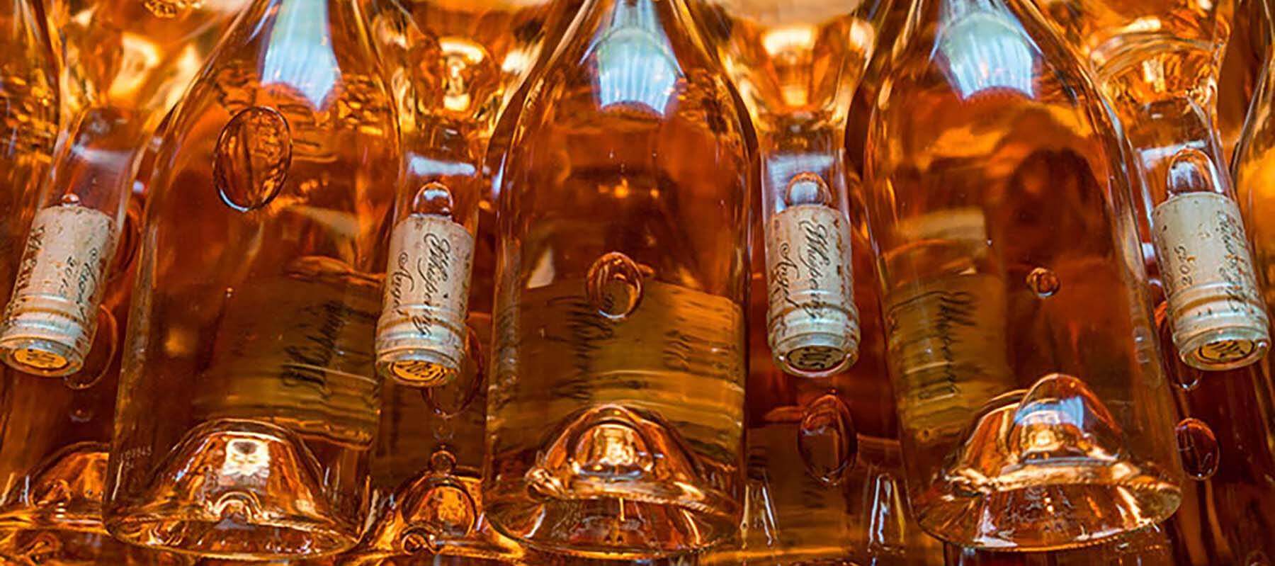 Close-up of several upside-down wine bottles with golden caps and amber liquid. The bottles are stacked horizontally, creating a warm, glowing effect as light passes through them, highlighting the texture and bubbles on the glass.