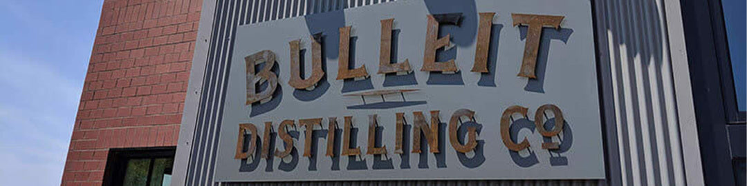 Sign reading Bulleit Distilling Co mounted on a brick and metal building exterior under a blue sky.