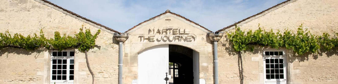 Facade of a historic stone building with green vines and two windows. The entrance features an arched sign that reads Martell The Journey.