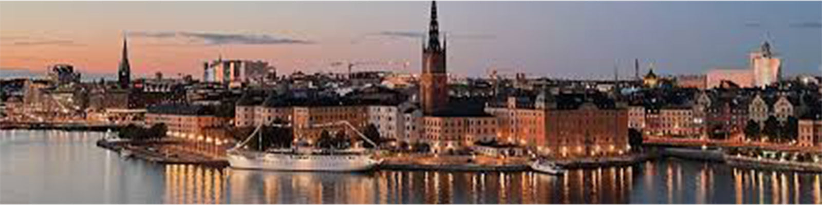 A panoramic view of a cityscape by the water at sunset, featuring historic buildings and a tall church steeple. Warm lights reflect on the water, and a white ship is docked at the quay.