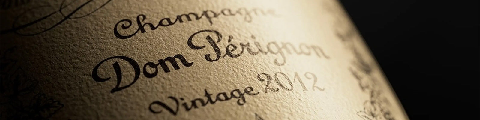 Close-up of a Dom Pérignon champagne bottle label, featuring elegant cursive text Champagne Dom Pérignon Vintage 2012 on a textured beige background, with intricate design patterns surrounding the text.