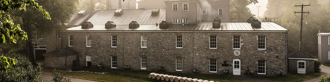 Historic stone building surrounded by trees, featuring a metal roof and multiple windows. Several barrels are lined up by the road in front of the building, with early morning fog in the background.