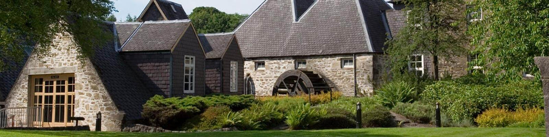 A scenic view of a traditional stone building with slate roofs and a large central water wheel, surrounded by lush greenery and well-maintained gardens.