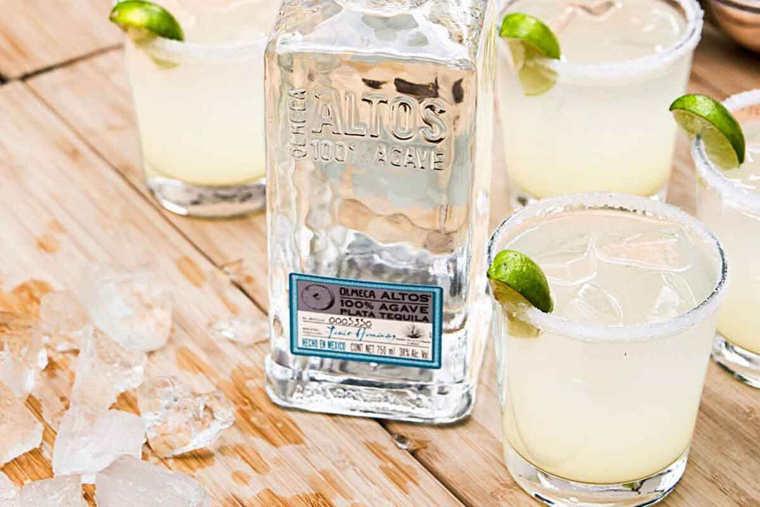A bottle of Olmeca Altos tequila is surrounded by four margaritas on a wooden surface. Each glass is garnished with a lime wedge and has a salted rim. Ice cubes are scattered nearby.