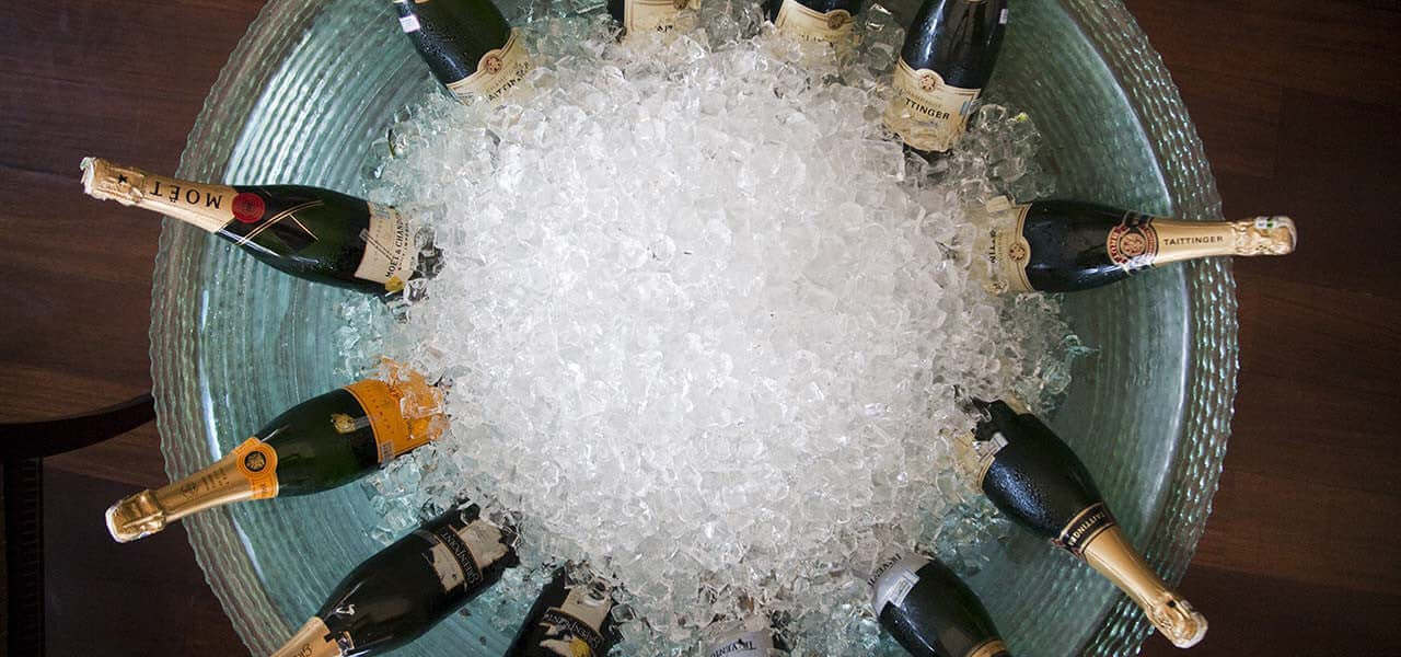 A large round glass bowl filled with ice holds various bottles of champagne, arranged in a circular pattern with their necks pointing outward. The bowl is placed on a wooden surface.