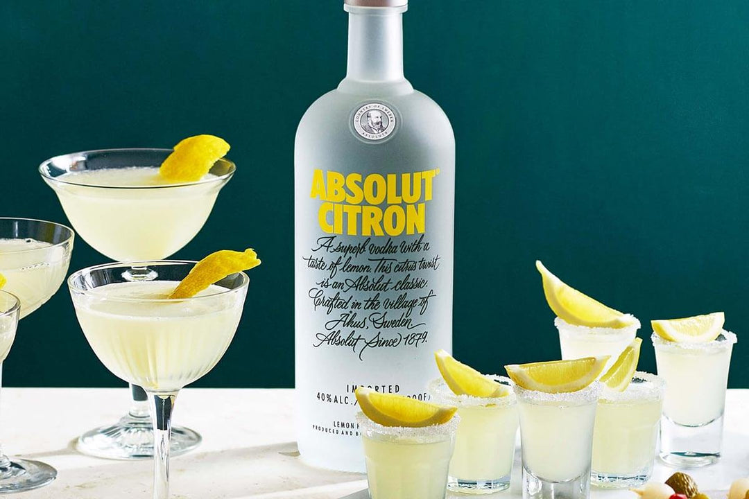 A bottle of Absolut Citron vodka stands surrounded by cocktails garnished with lemon twists. The glasses are elegantly arranged, showcasing the clear, lemon-infused drinks against a dark green background.