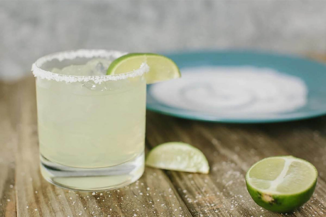 A glass of margarita with a salted rim and lime wedge sits on a wooden table. A cut lime and a blue dish with salt are in the background.