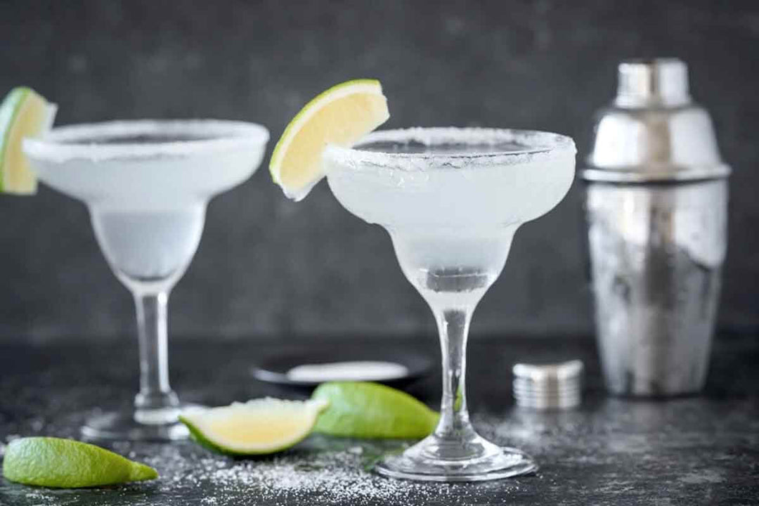Two margarita glasses with salted rims and lime wedges, placed on a dark surface. A metal cocktail shaker and additional lime slices are in the background.