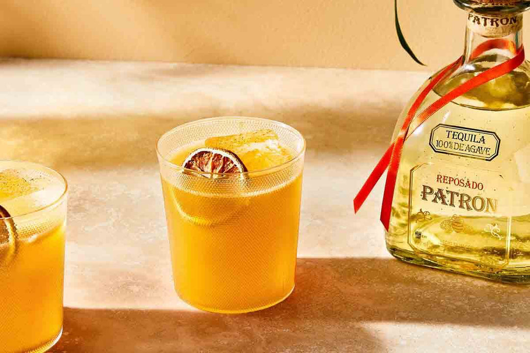Two glasses of an orange cocktail with ice and garnished with dried citrus slices sit on a table beside a bottle of Patrón Reposado Tequila. Sunlight casts a warm glow over the scene.