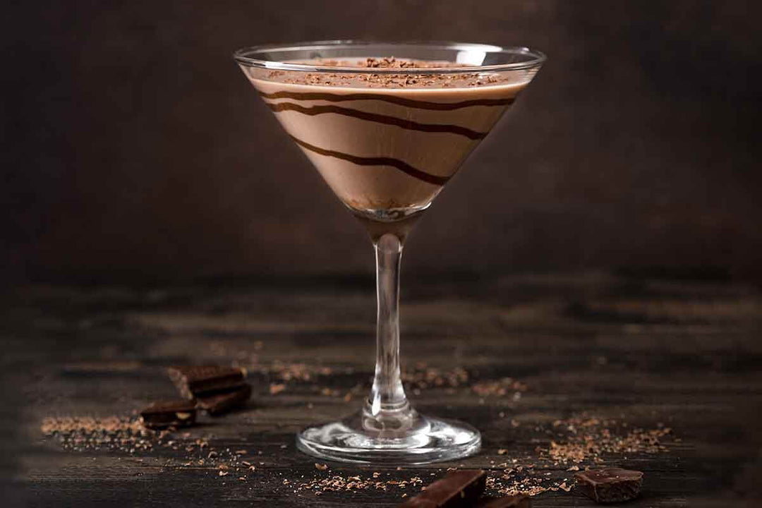 A martini glass filled with a creamy chocolate cocktail, drizzled with chocolate syrup, and topped with chocolate shavings. Dark chocolate pieces and cocoa powder are scattered on the dark wooden surface beneath the glass.