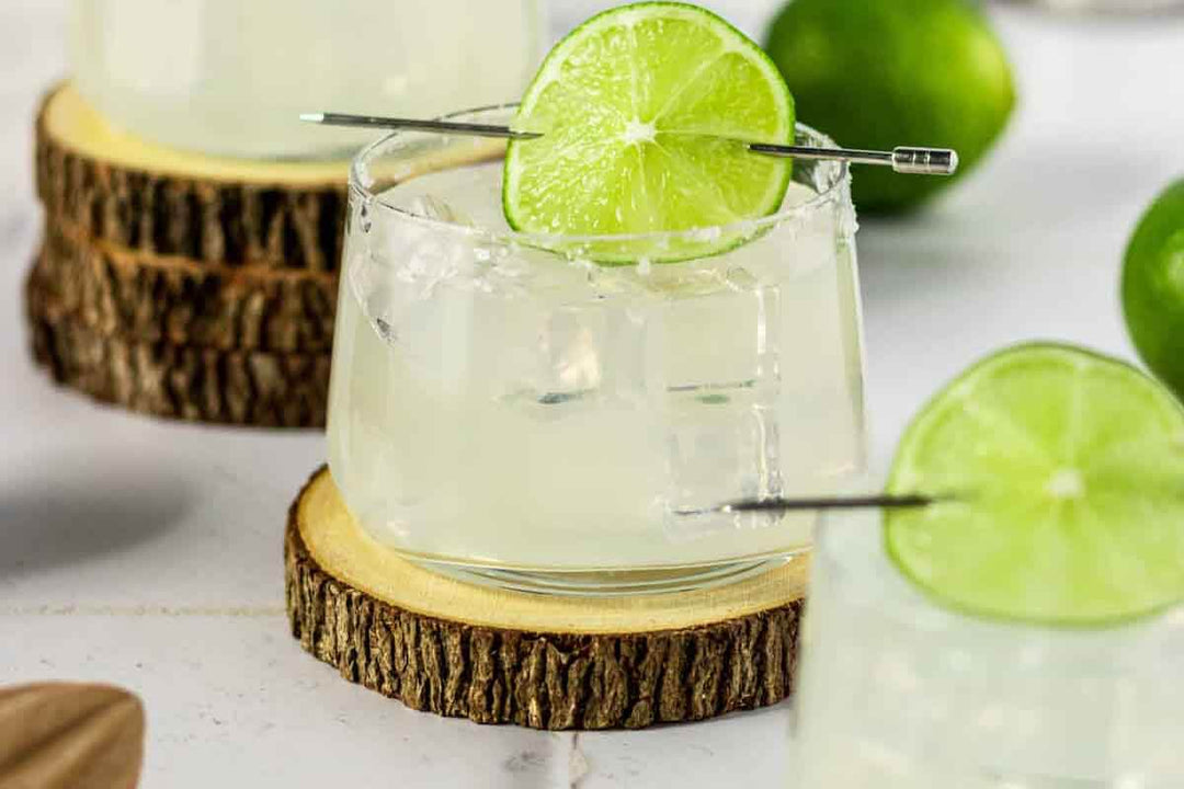 A glass of margarita with salt rim, garnished with a lime slice, placed on a wooden coaster. Additional limes and similar glasses in the background on a white surface.
