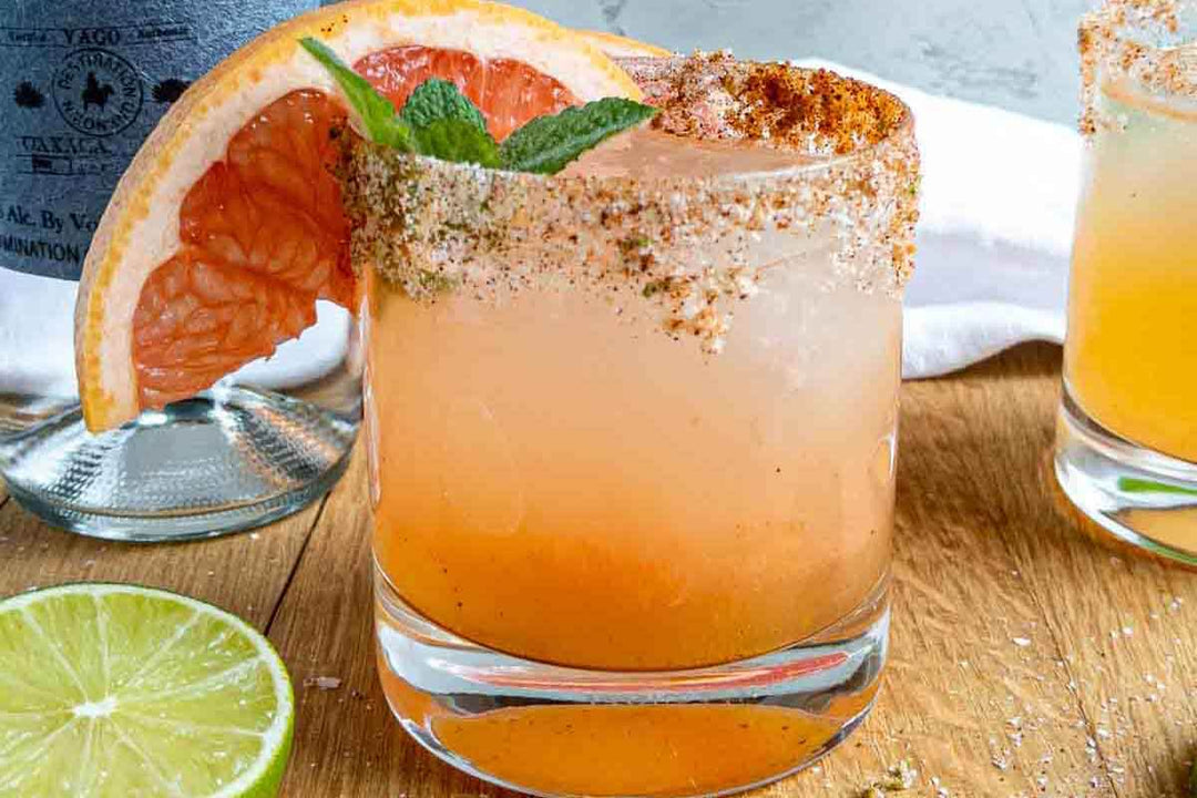 A refreshing cocktail with a slice of grapefruit and a sprig of mint garnish in a glass with a spiced rim. Another similar drink is visible in the background on a wooden surface, next to a halved lime.