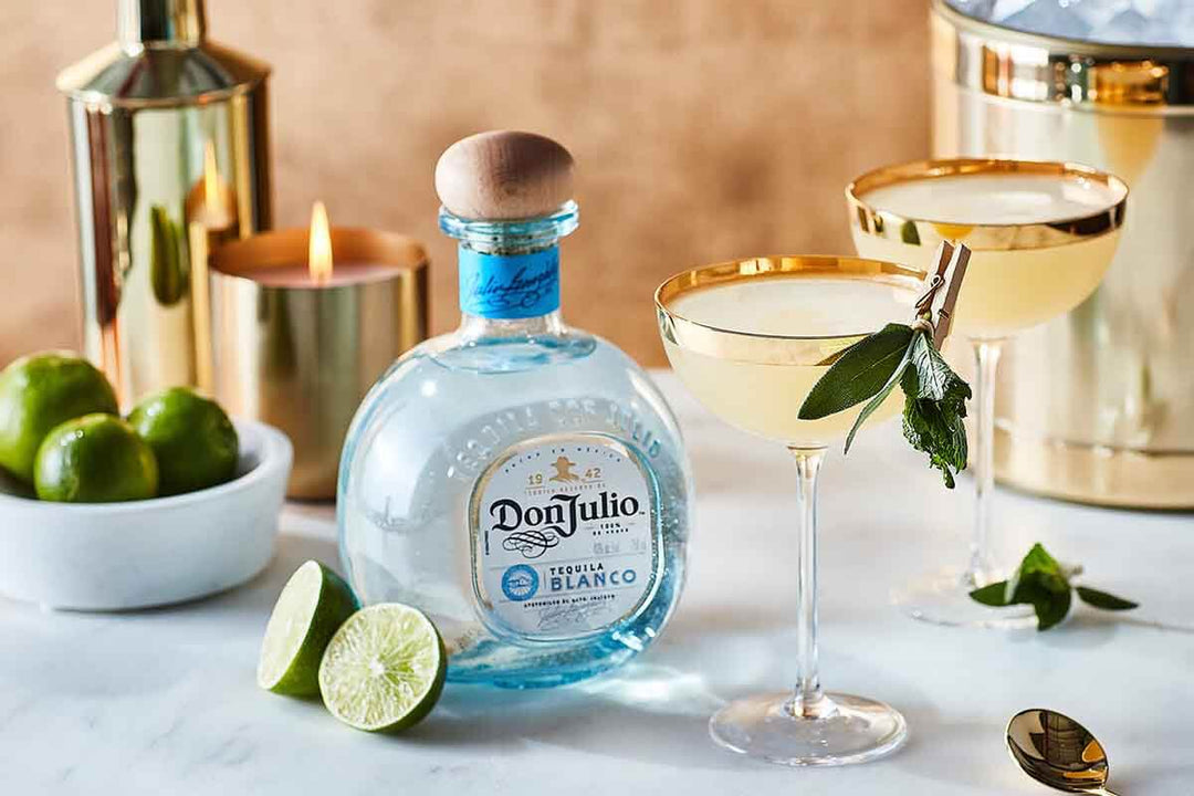 A bottle of Don Julio Tequila Blanco sits beside two cocktails garnished with herbs. Limes, a lit candle, and a shiny cocktail shaker are in the background on a marble surface.