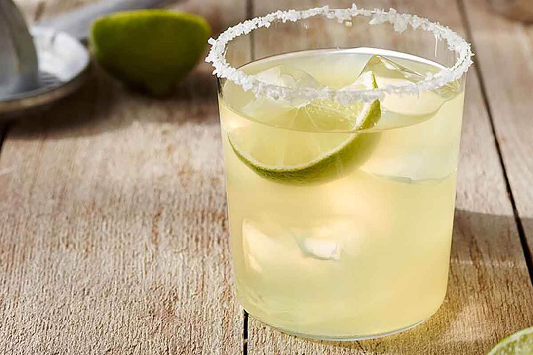 A glass of margarita with a salted rim and ice cubes sits on a wooden table. A lime wedge is inside the drink. In the background, a lime and a juicer are slightly out of focus.