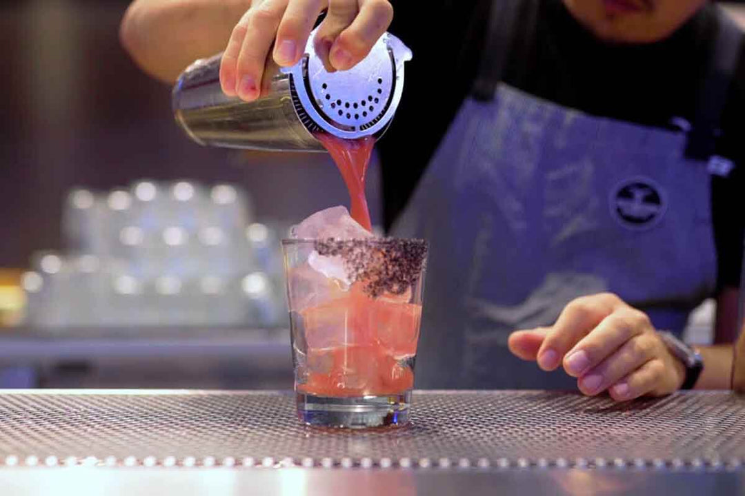 A bartender pours a pink cocktail from a metal shaker into a glass filled with ice. The bartender wears a dark apron, and the scene is set on a metal counter with an out-of-focus background.