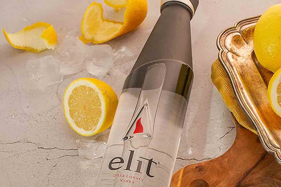 A bottle of Elit Ultra Luxury Vodka lies on a textured surface surrounded by lemon slices and peel. Ice cubes are scattered around, and a brass tray is visible in the background, adding an elegant touch to the scene.