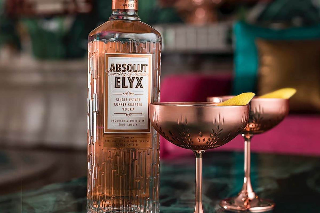 A bottle of Absolut Elyx vodka stands on a table next to two copper martini glasses filled with a clear liquid and garnished with lemon twists. The background features a blurred, colorful interior setting.