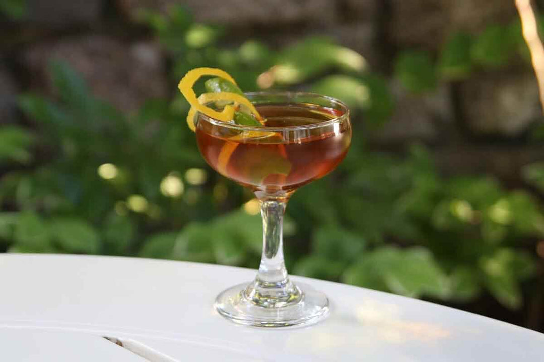 A cocktail in a coupe glass garnished with a citrus twist. The drink is placed on a white table with a blurred background of green foliage, creating an inviting and refreshing atmosphere.