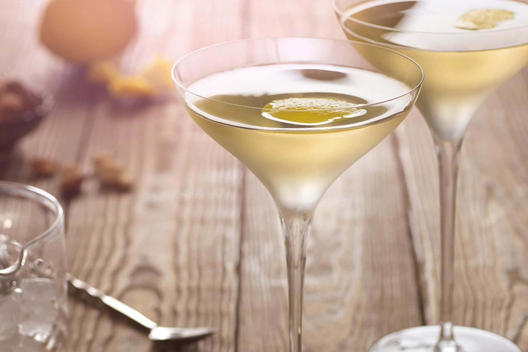 Two elegant cocktail glasses filled with a light golden drink, garnished with a lemon twist. The glasses are on a wooden table with blurred lemons and nuts in the background, creating a warm and inviting ambiance.