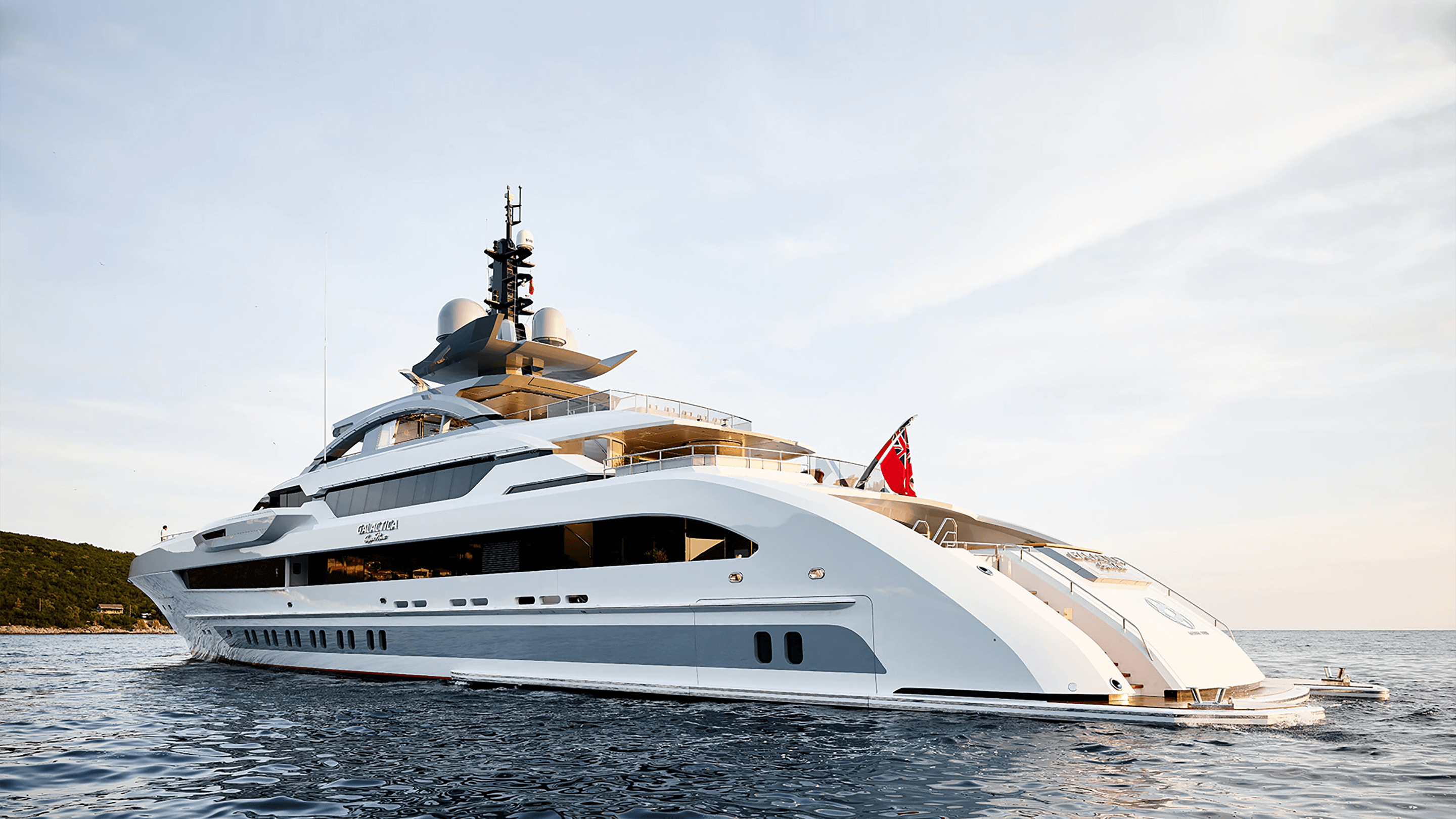 Luxury yacht sailing on calm waters with a sleek white and gray design. The vessel has multiple decks, large windows, and a flag at the stern. The backdrop features a green hillside under a clear sky with soft light.