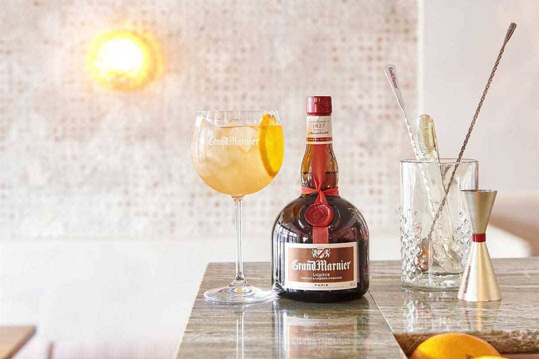 A bottle of Grand Marnier stands on a marble counter next to a cocktail glass filled with an orange drink, garnished with an orange slice. Nearby, a glass contains a spoon and strainer, with a jigger beside it. A bright light shines in the background.