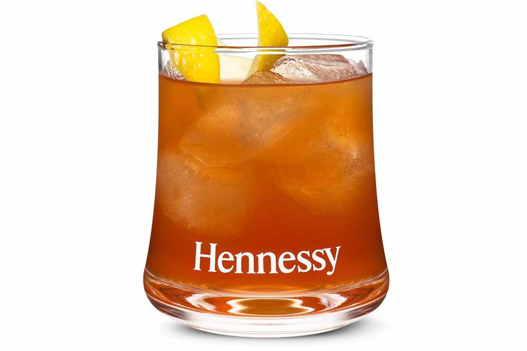 A glass of Hennessy cognac on ice, garnished with lemon twists, displaying a rich amber color. The word Hennessy is printed on the glass.