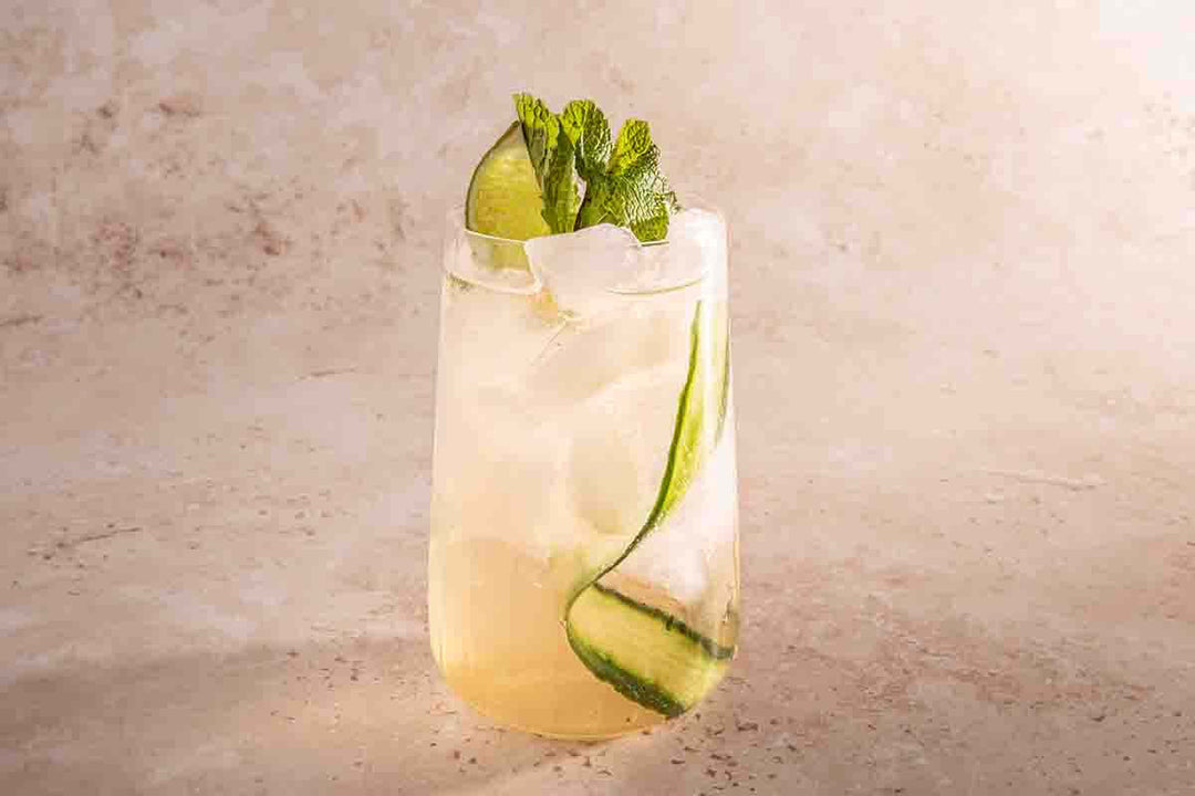 A tall glass filled with a refreshing cocktail, featuring ice cubes, lime wedges, cucumber slices, and mint leaves. The drink is set against a light, neutral background.