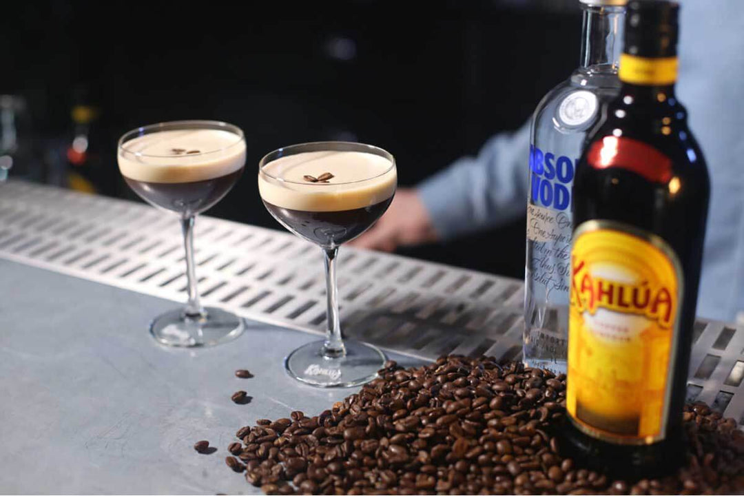 Two espresso martini cocktails in glasses sit on a bar counter beside coffee beans. A bottle of vodka and a bottle of Kahlúa are visible, suggesting key ingredients for the cocktails.