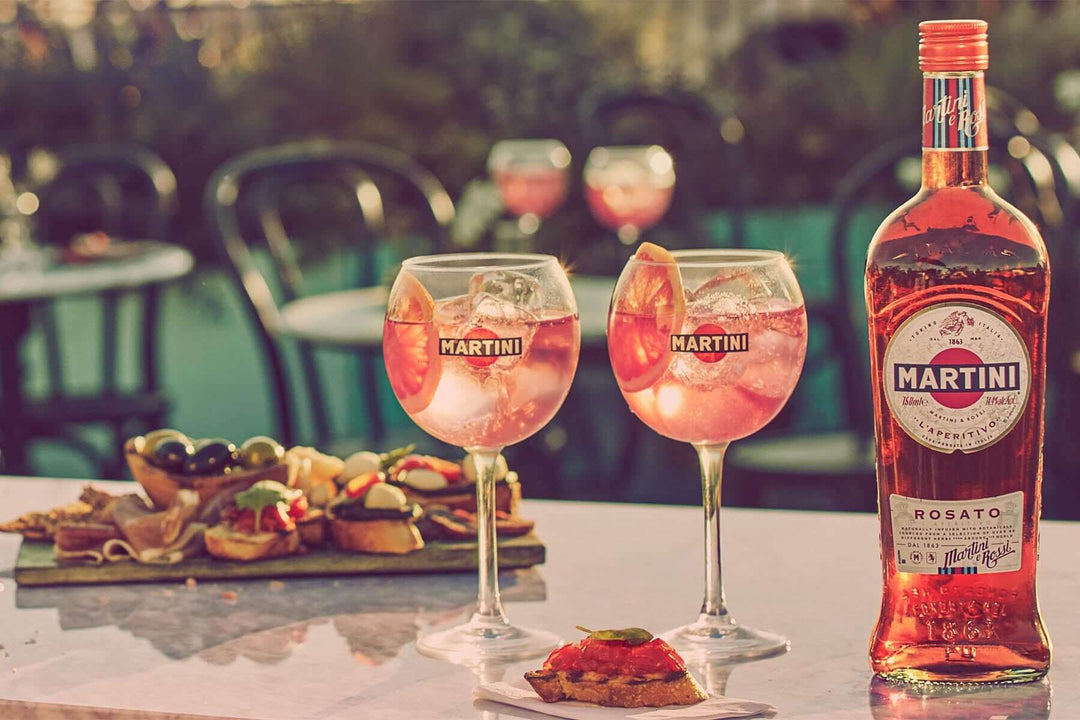 Two glasses of Martini Rosato with ice and citrus slices sit on a table next to a bottle of Martini Rosato. In the background, theres a plate with appetizers and outdoor seating with blurred chairs and tables.