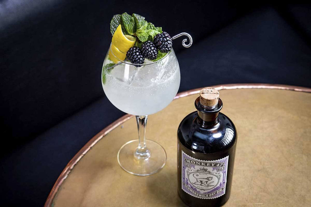A glass of gin and tonic garnished with lemon peel, blackberries, and mint sits on a round table. A bottle labeled Monkey 47 Schwarzwald Dry Gin is next to the glass. The background features a dark sofa.