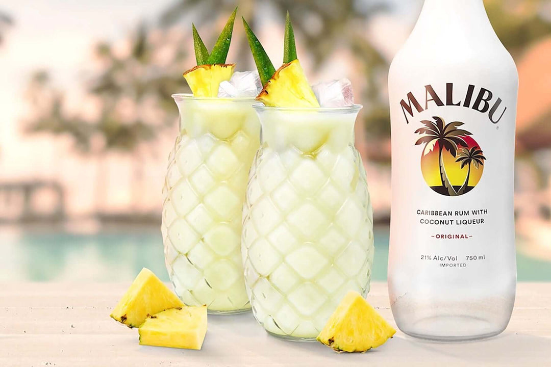 Two tropical drinks in pineapple-shaped glasses garnished with pineapple slices sit beside a bottle of Malibu rum. Pineapple wedges are scattered on the table, and a blurred poolside background creates a summery atmosphere.