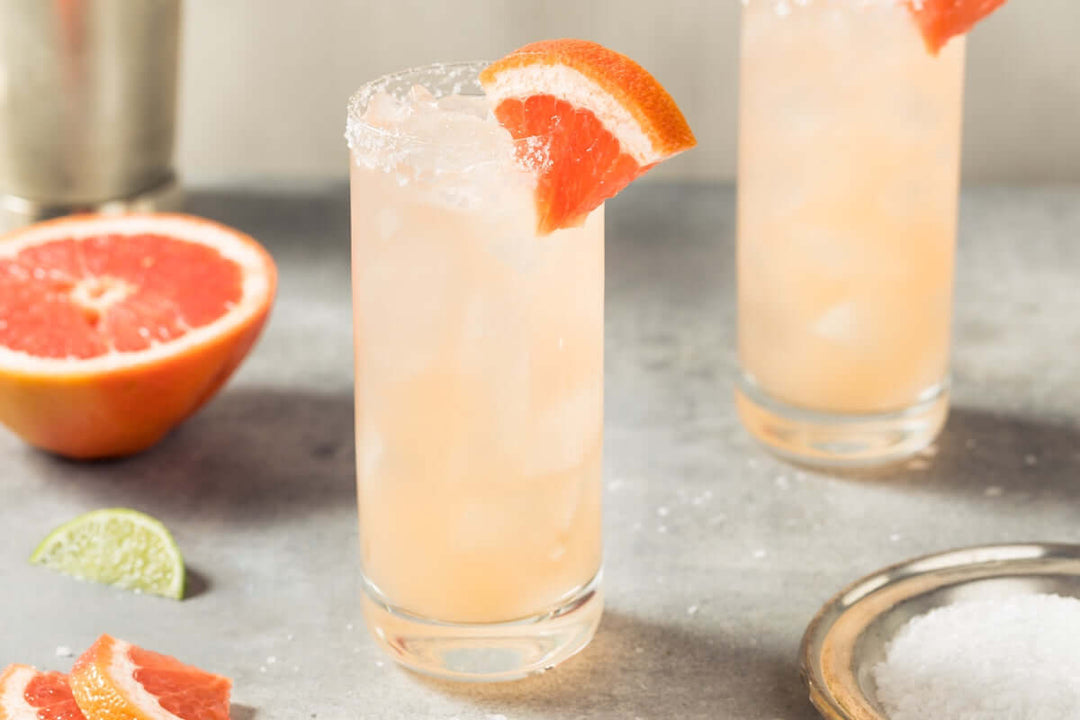A refreshing cocktail served in a tall glass with a salted rim, garnished with a grapefruit slice. Another similar drink is blurred in the background. Fresh grapefruit and lime slices are scattered on the table.