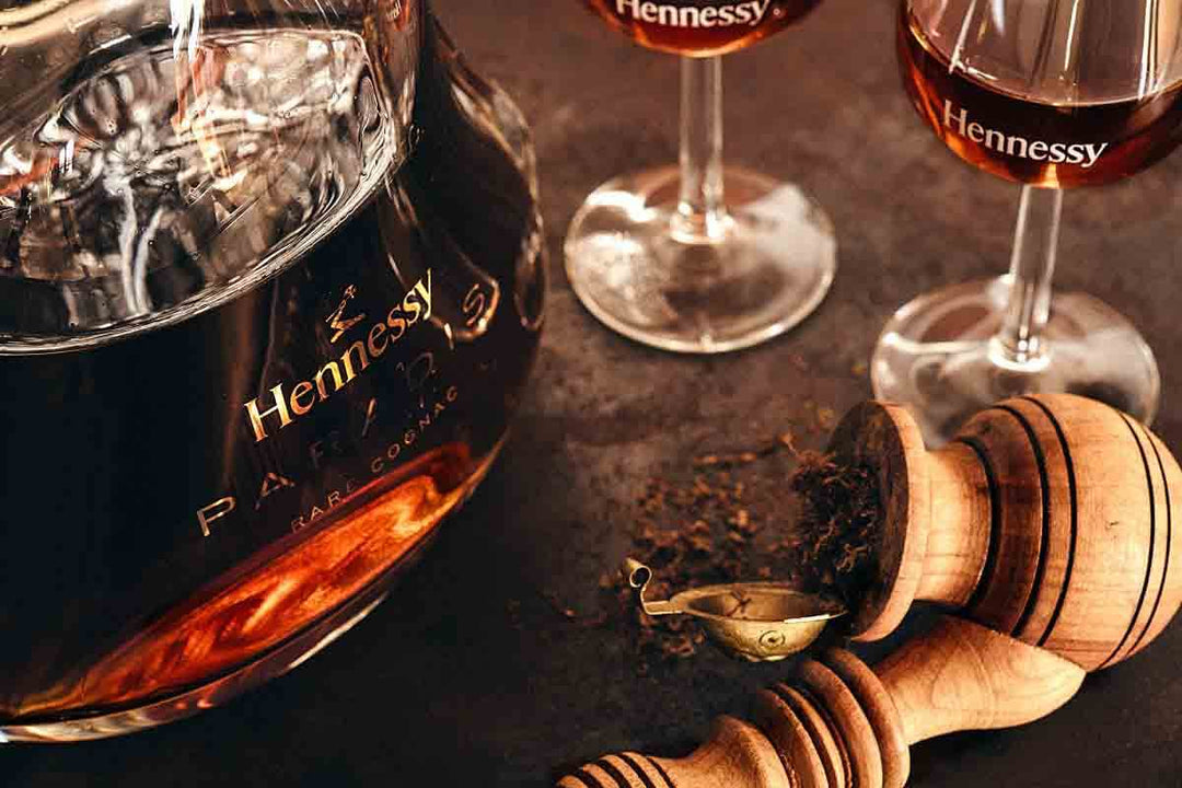 A close-up of a bottle of Hennessy cognac with two filled glasses next to it on a table. A wooden muddler and crushed spices are nearby, creating a warm and inviting atmosphere.