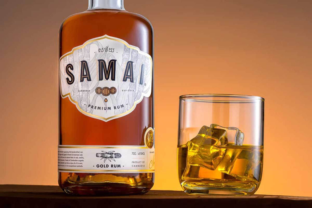 A bottle of Samai Premium Gold Rum stands next to a glass filled with rum and ice cubes. The background is a warm, golden hue, enhancing the rich color of the rum.
