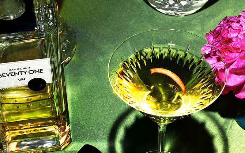 A bottle of Seventy One gin sits on a green surface next to a crystal glass with a served drink and a pink flower. The gin bottle has a black and white label, and the drink in the glass has a twist of citrus peel.