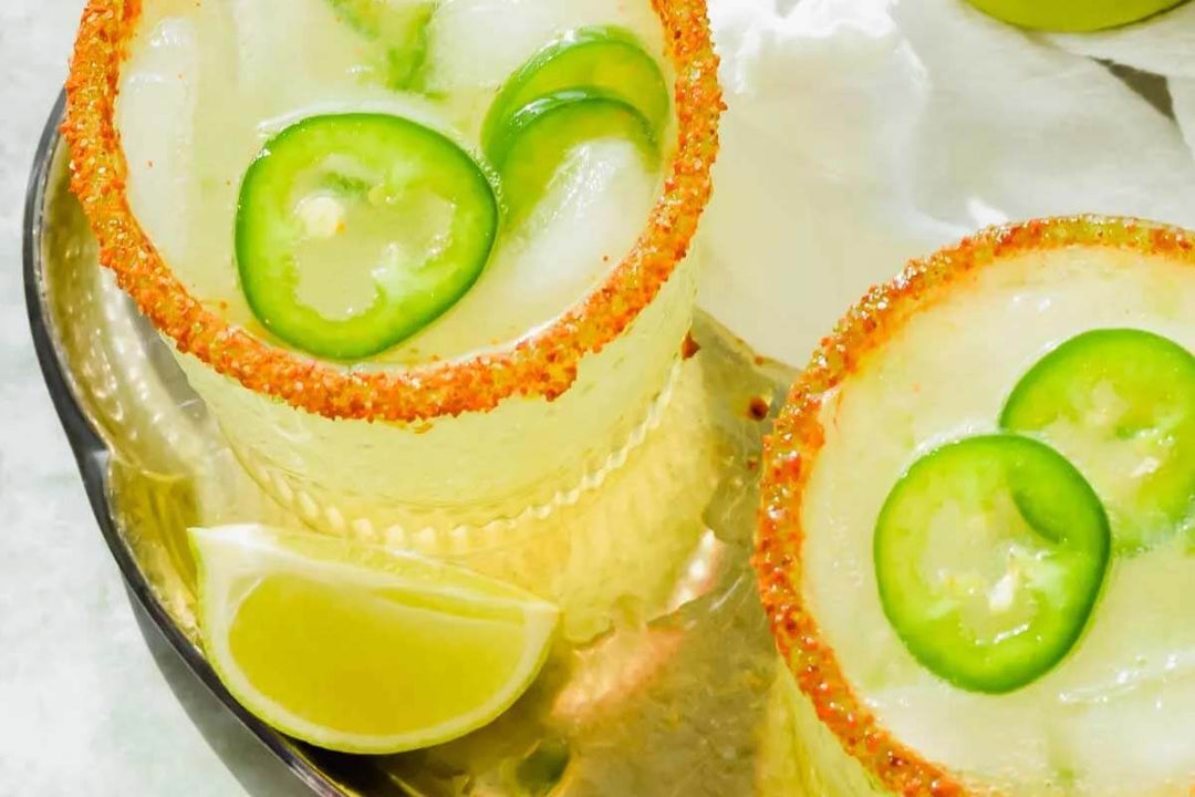 Two glasses of spicy margarita with ice and jalapeño slices, rimmed with chili powder. A lime wedge sits nearby on a metal tray.