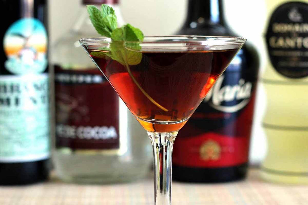 A martini glass filled with a dark cocktail, garnished with a mint sprig, is centered in the foreground. Blurry liquor bottles are visible in the background, suggesting a bar setting.