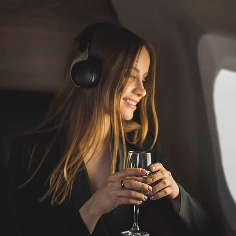 A person with long hair, wearing headphones, smiles while holding a glass. They are seated by a window, possibly in an airplane, admiring the view outside.