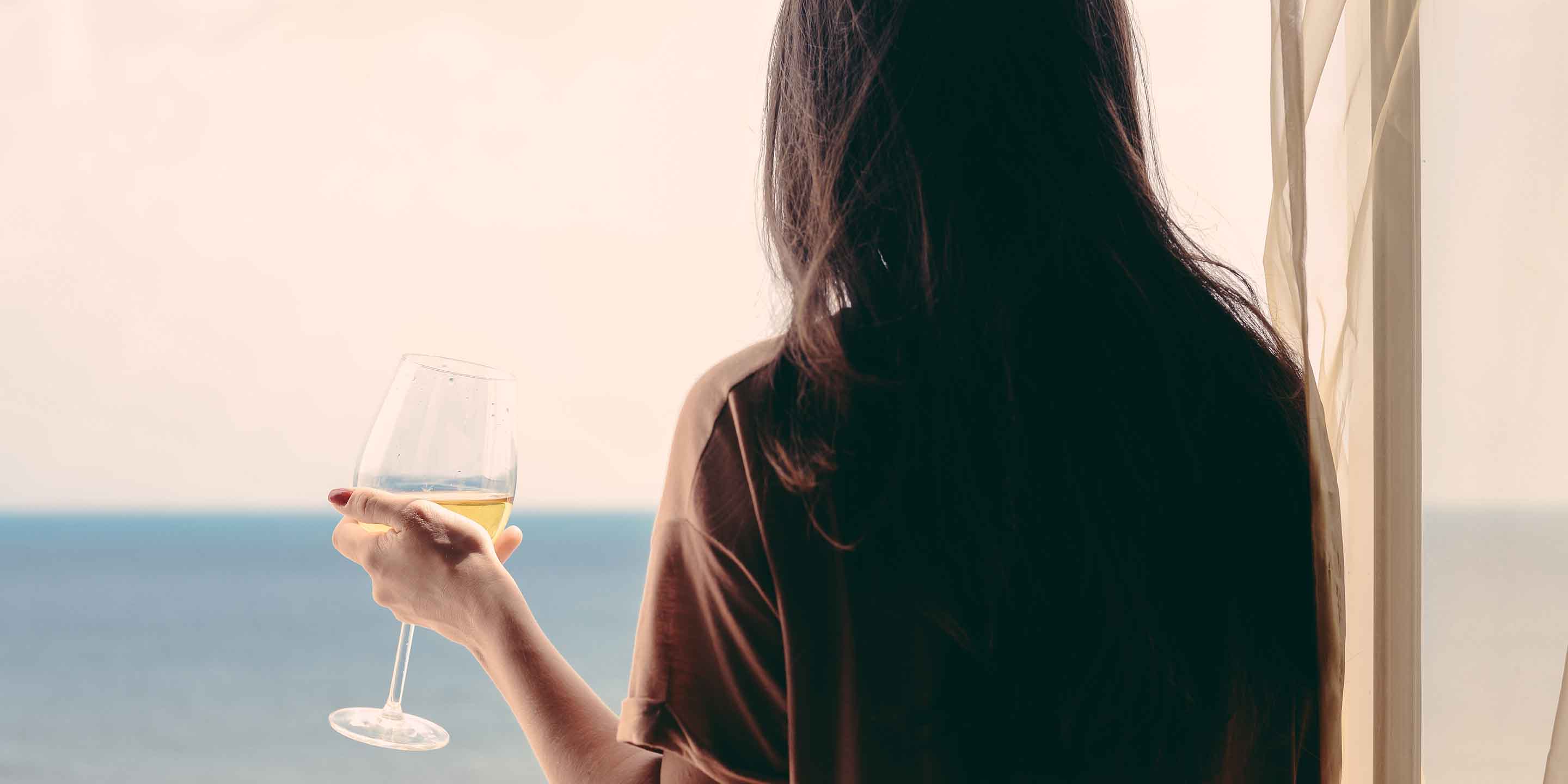 A woman with long dark hair stands by a window, holding a glass of white wine. She gazes out at a serene view of the ocean on a clear day. The atmosphere is calm and reflective.