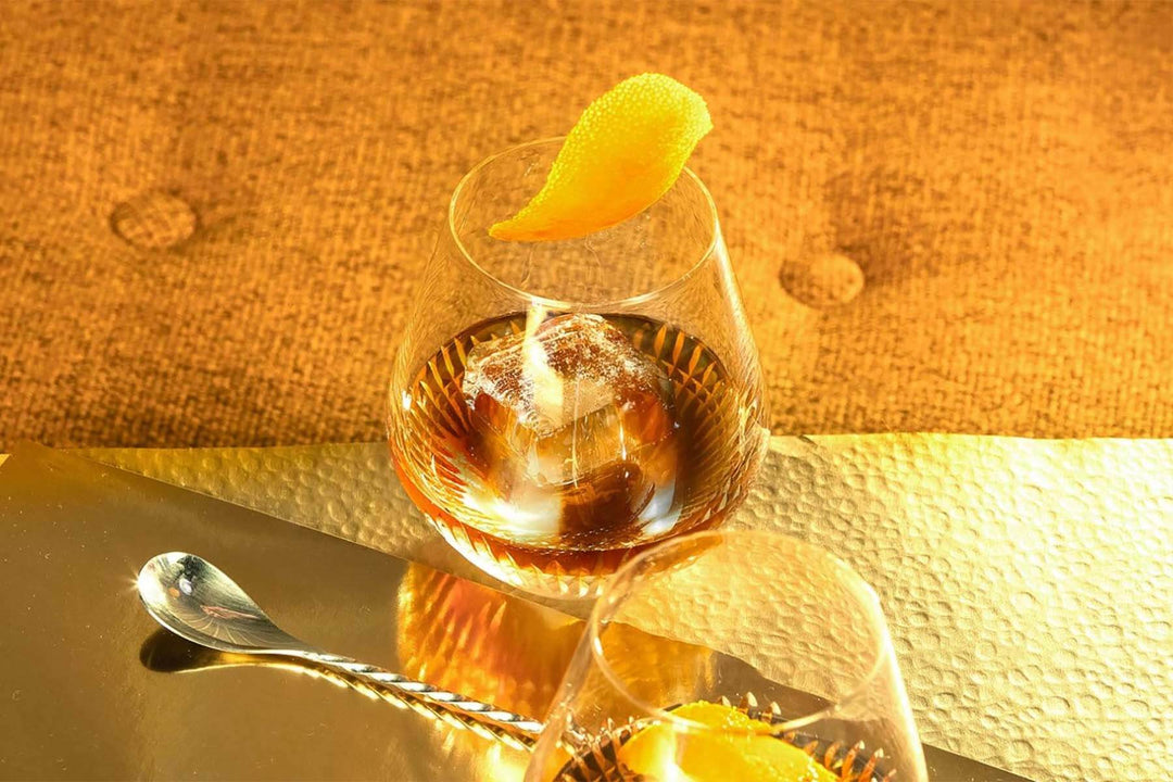 A glass of whiskey with a large ice cube and an orange peel garnish sits on a reflective surface. A twisted metal spoon is nearby, and the background is a warm, textured fabric. The scene is bathed in a golden light.