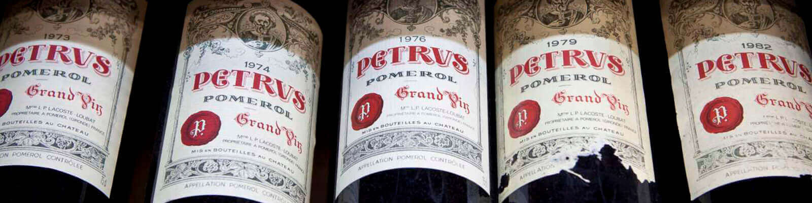 Close-up of five bottles of Petrus Pomerol wine, displaying labels from vintages 1974, 1975, 1976, 1978, and 1979. Each label features intricate designs with Grand Vin prominently printed in red.