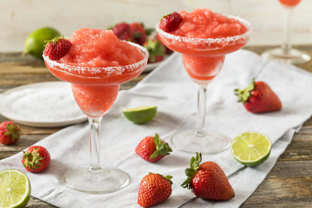 Two glasses of frozen strawberry margaritas sit on a cloth, garnished with strawberries and rimmed with salt. Surrounding them are fresh strawberries and lime halves on a wooden surface.